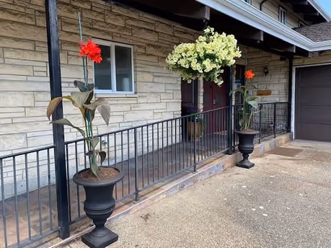 Exterior view of a building with a stone facade, a covered porch with a black metal railing, and three large black planters containing flowers. Two planters have red flowers and one has white flowers. The entrance door is visible in the background.