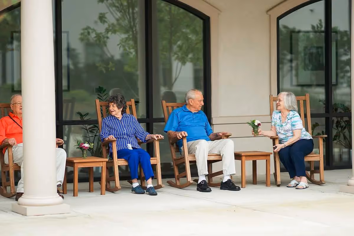 Four elderly people sitting on wooden rocking chairs on a covered patio outside a building. Two small wooden tables with flower arrangements are placed between the chairs. The group appears to be engaged in conversation and enjoying the outdoor setting.