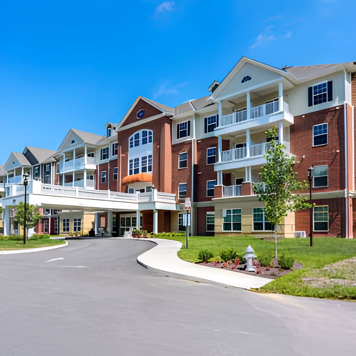 Exterior view of a multi-story senior living facility building with red brick and white accents, featuring balconies, large windows, a covered entrance driveway, and a well-maintained lawn with small trees and shrubs under a clear blue sky.