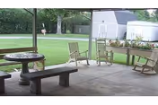 Covered patio with a round table, concrete benches, wooden rocking chairs, planters, and a grassy yard with storage sheds beyond.