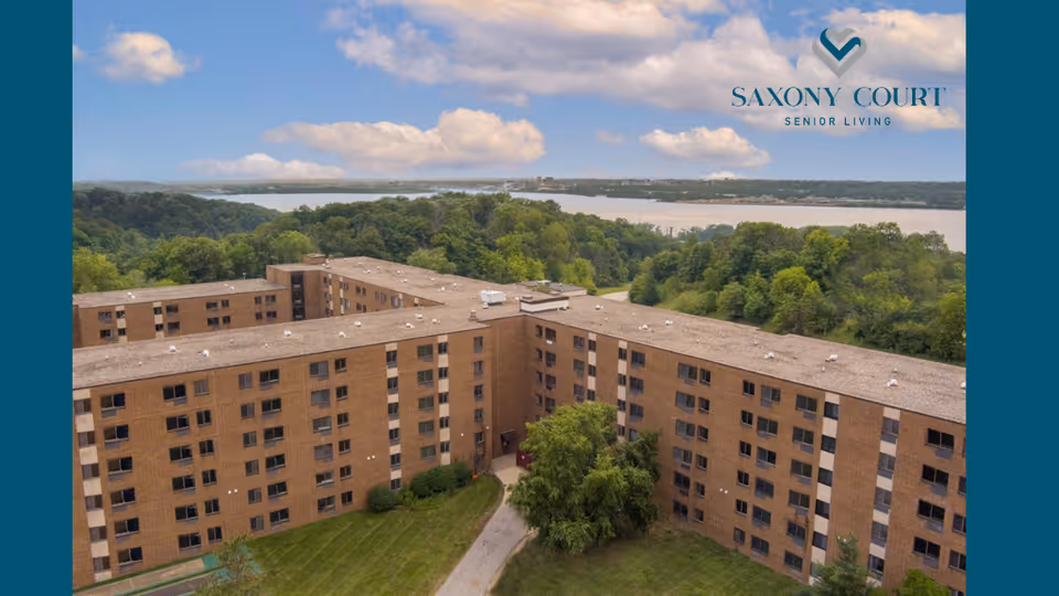 Aerial view of a large, multi-story brick senior living facility named Saxony Court Senior Living, surrounded by green trees and overlooking a body of water under a partly cloudy sky.
