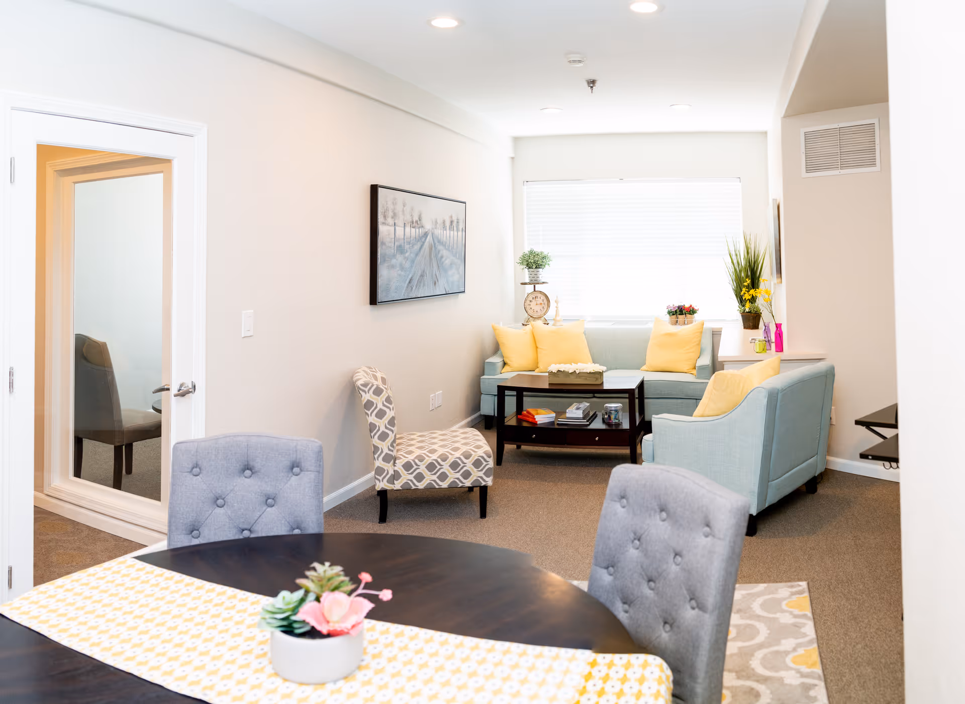 Bright living room and dining area with light blue sofas, yellow pillows, a coffee table, and a round dining table with gray tufted chairs.