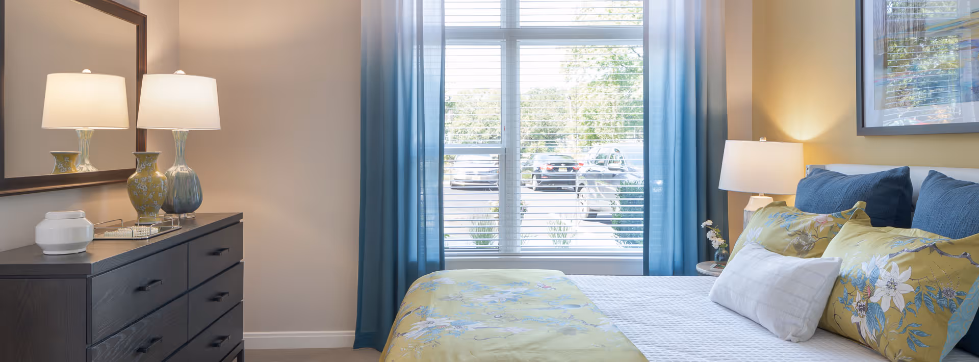 A bright and cozy bedroom with a large window covered by blue curtains, a bed with floral and solid blue pillows and a yellow floral bedspread, a dark wooden dresser with two decorative lamps and a mirror above it, and a framed picture on the wall.