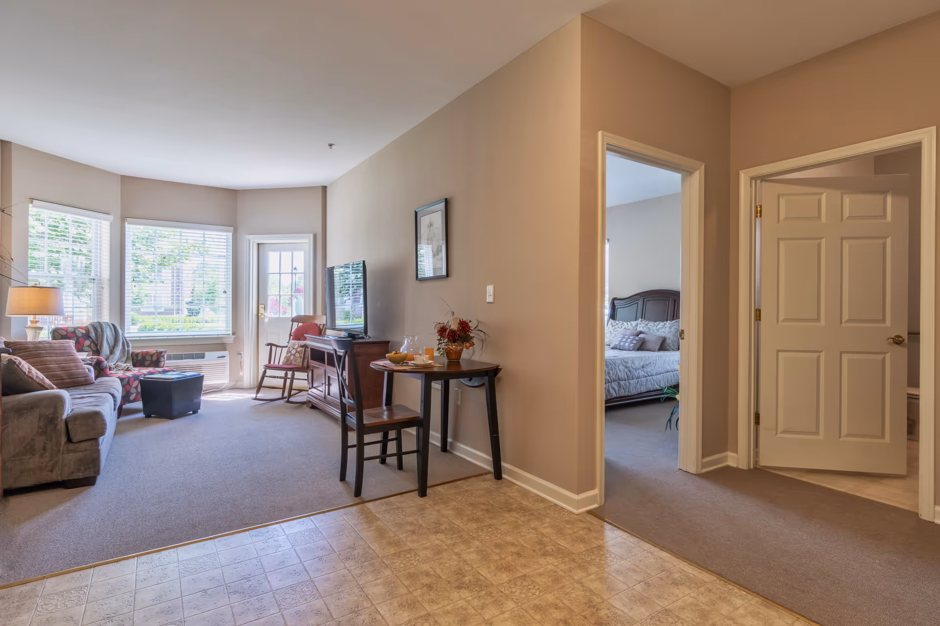 Sunlit living room with a sofa, armchair, TV, small dining table and a doorway leading to a bedroom.