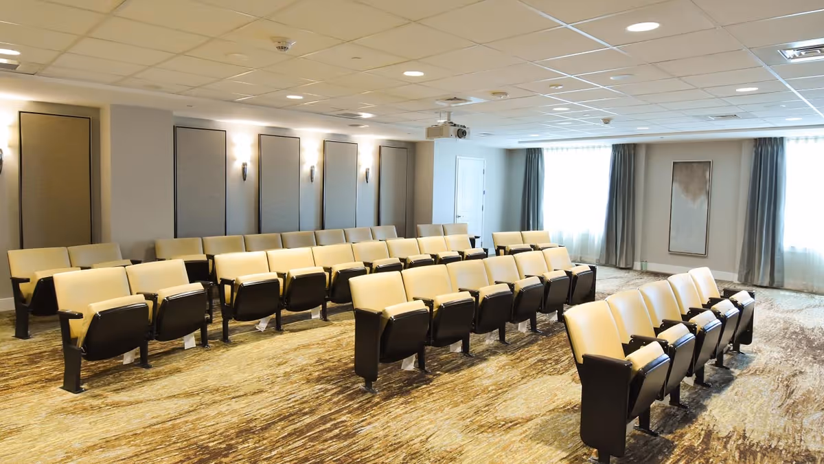 A well-lit meeting or presentation room with rows of beige cushioned chairs with black armrests arranged facing the front. The room has a patterned carpet, light-colored walls with vertical rectangular panels, wall sconces, and two large windows with gray curtains allowing natural light to enter. A ceiling-mounted projector is visible near the front of the room.