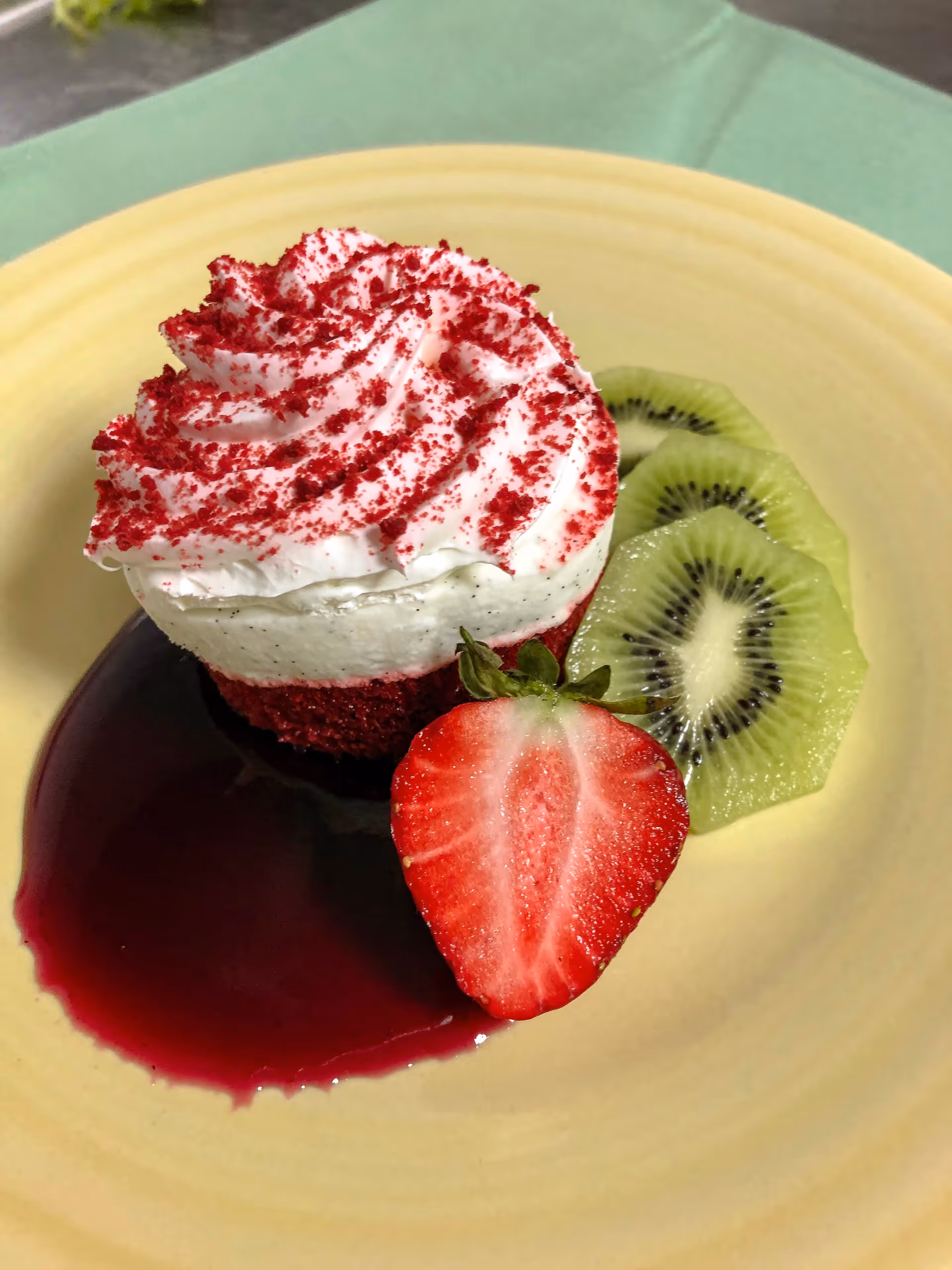 A dessert plate featuring a red velvet cupcake topped with white frosting and red sprinkles, accompanied by a sliced strawberry, two slices of kiwi, and a pool of dark red sauce on a yellow plate with a green tablecloth underneath.
