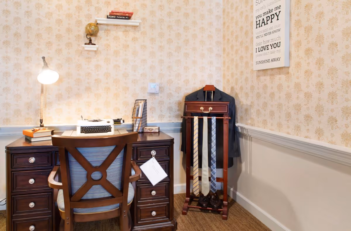 A small vintage-style desk and chair in a patterned wallpapered room with a lamp, typewriter, books, and a valet stand holding a suit and ties.