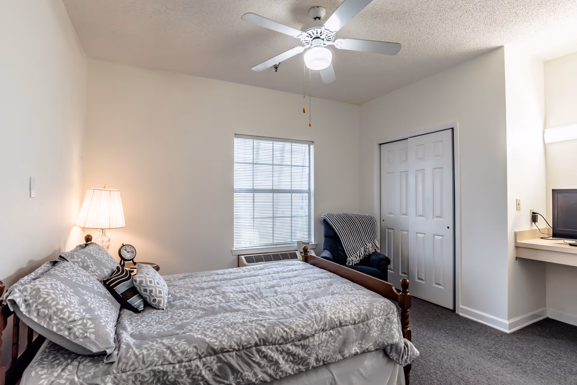 Bright bedroom with a made bed, bedside lamp and clock, window, armchair, closet, ceiling fan and small desk area.