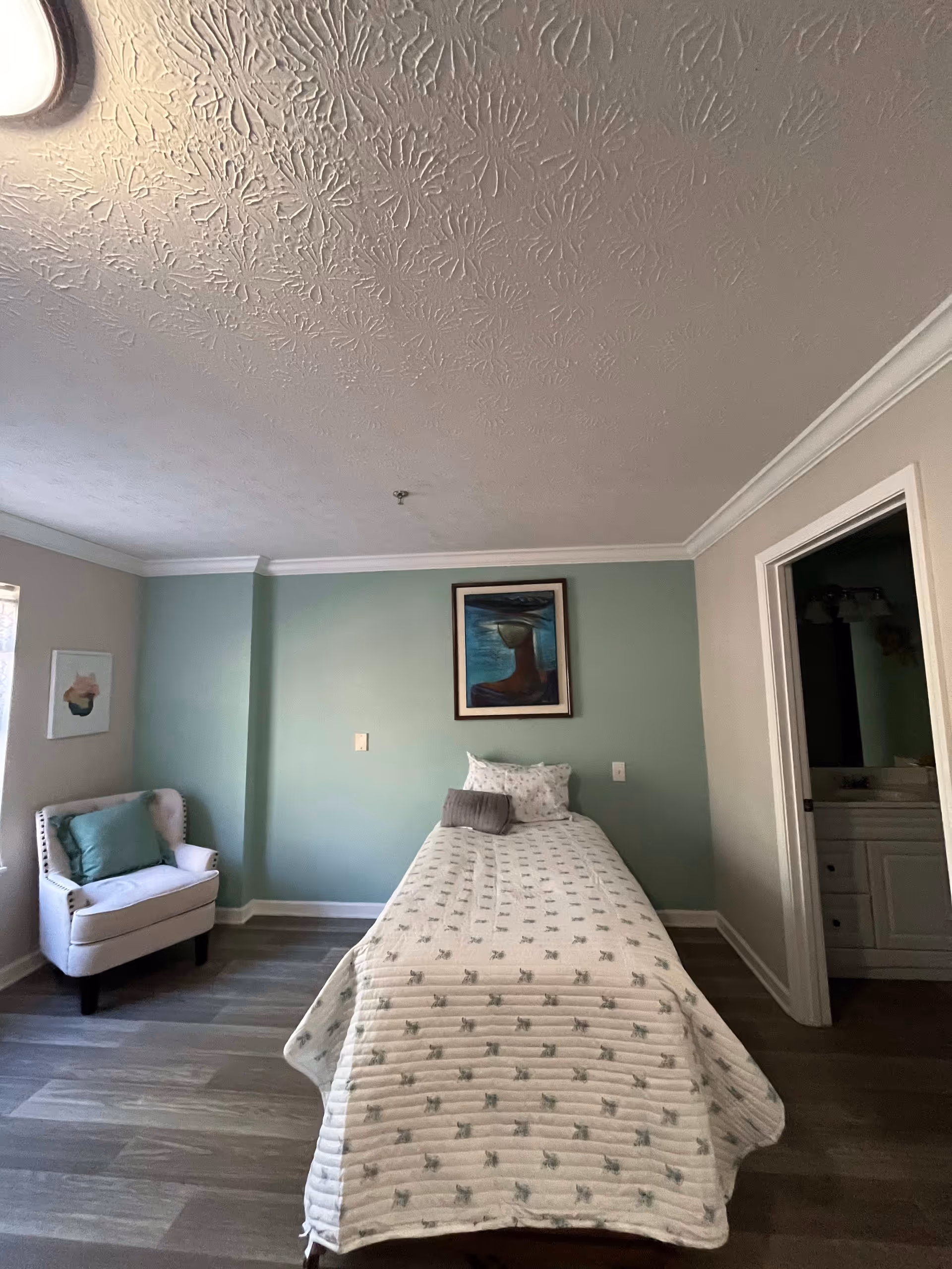 A simple bedroom with a single bed covered in a white quilt with a subtle pattern. There are two pillows on the bed. A framed painting hangs on the light green wall behind the bed. To the left, there is a white armchair with a green cushion near a window. The floor is wood, and to the right, there is an open doorway leading to a bathroom with a white vanity and mirror.