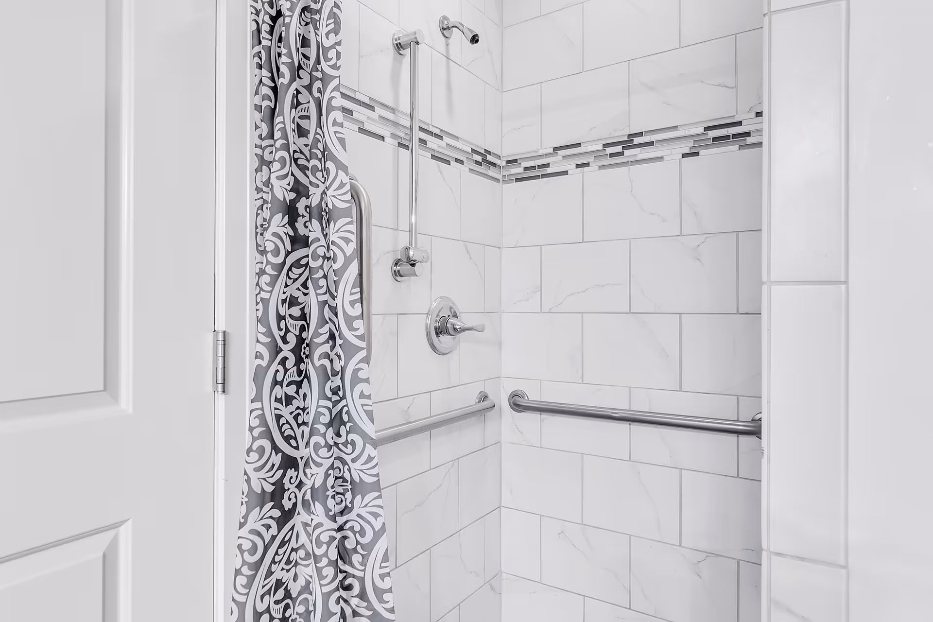 A tiled shower area with white and gray marble-patterned tiles and a decorative horizontal strip of smaller tiles. The shower has a silver showerhead, two silver grab bars on the walls, and a gray and white patterned shower curtain partially drawn to the side.