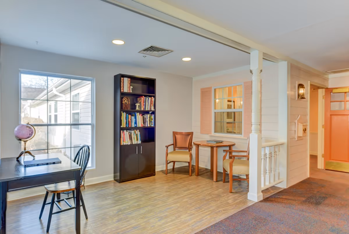 A cozy interior room with a large window letting in natural light. There is a black desk with a globe and a chair near the window. A tall black bookshelf filled with books stands against the wall. Two wooden chairs with cushions are placed around a small round wooden table. The room has light-colored walls, wood flooring, and a carpeted area leading to a hallway with an orange door.