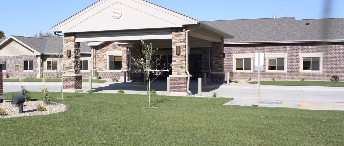 Exterior view of a single-story brick building with a covered entrance supported by stone pillars, surrounded by a well-maintained lawn and small trees under a clear sky.