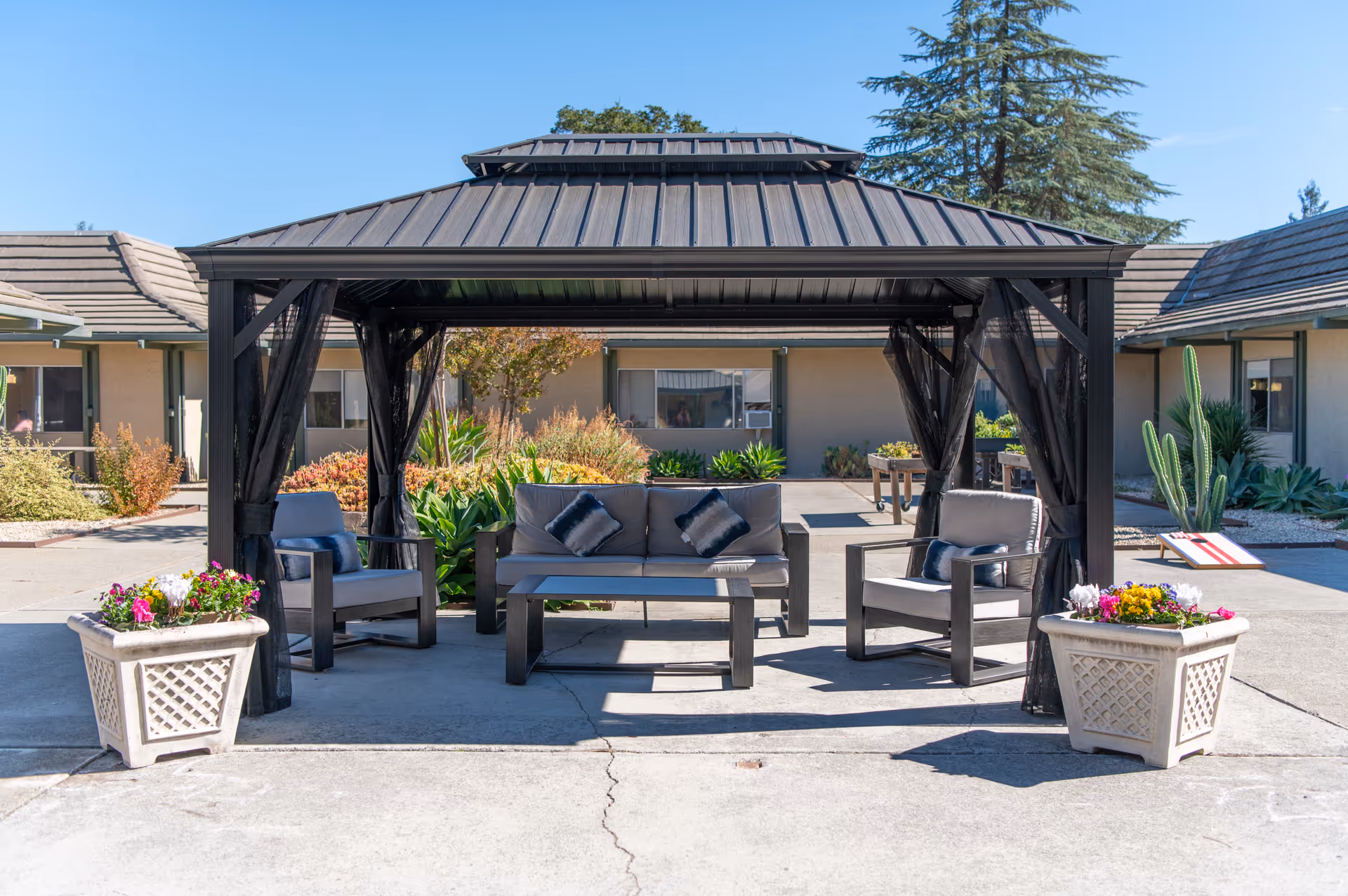 Outdoor courtyard featuring a metal gazebo with patio seating, planters, and a single-story building in the background.