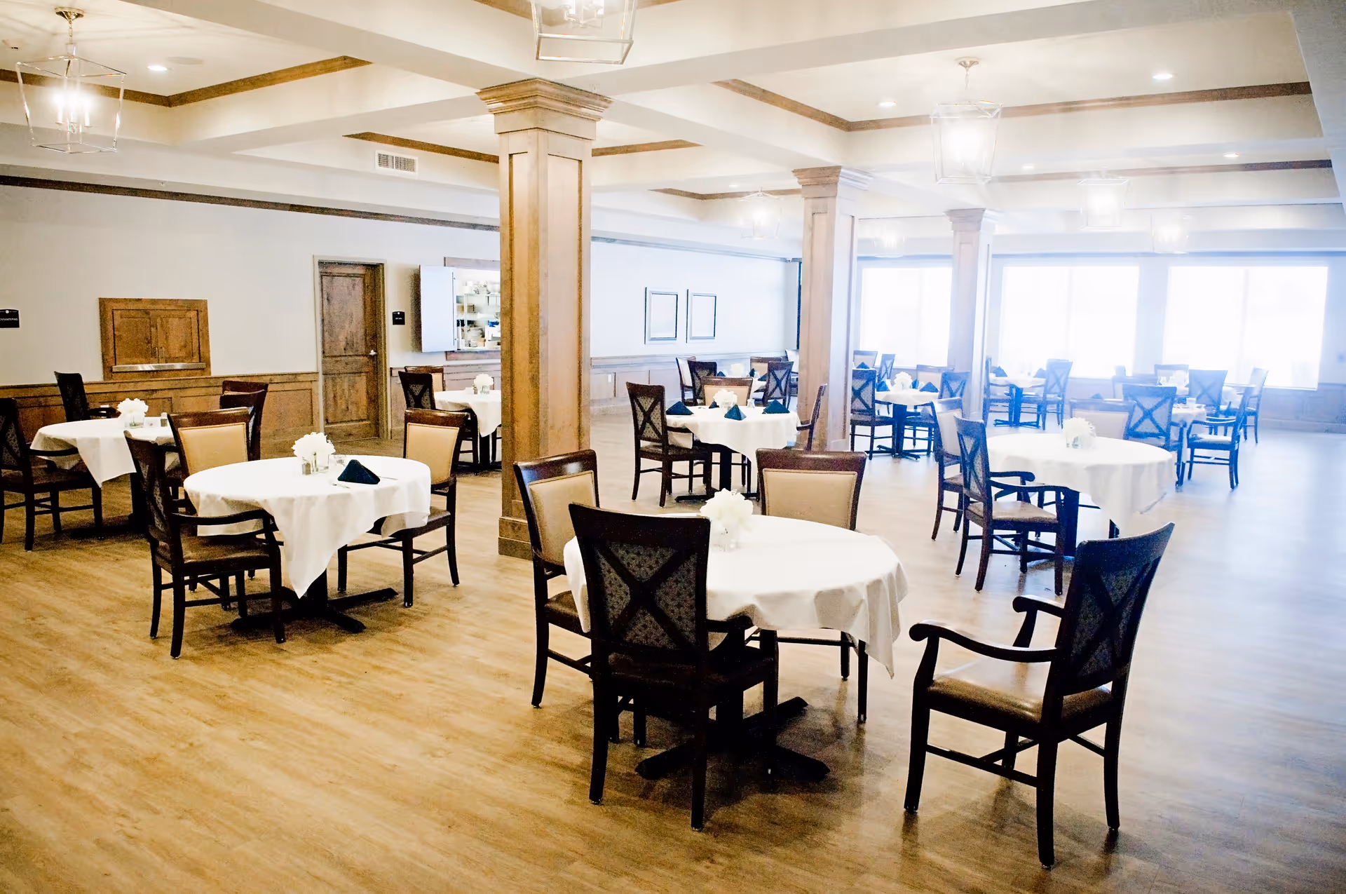 A spacious dining room with multiple round tables covered with white tablecloths, each set with chairs and small floral centerpieces. The room features wooden flooring, wooden columns, and large windows allowing natural light to fill the space.