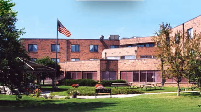 Brick senior living facility with a manicured lawn, benches, and an American flag.