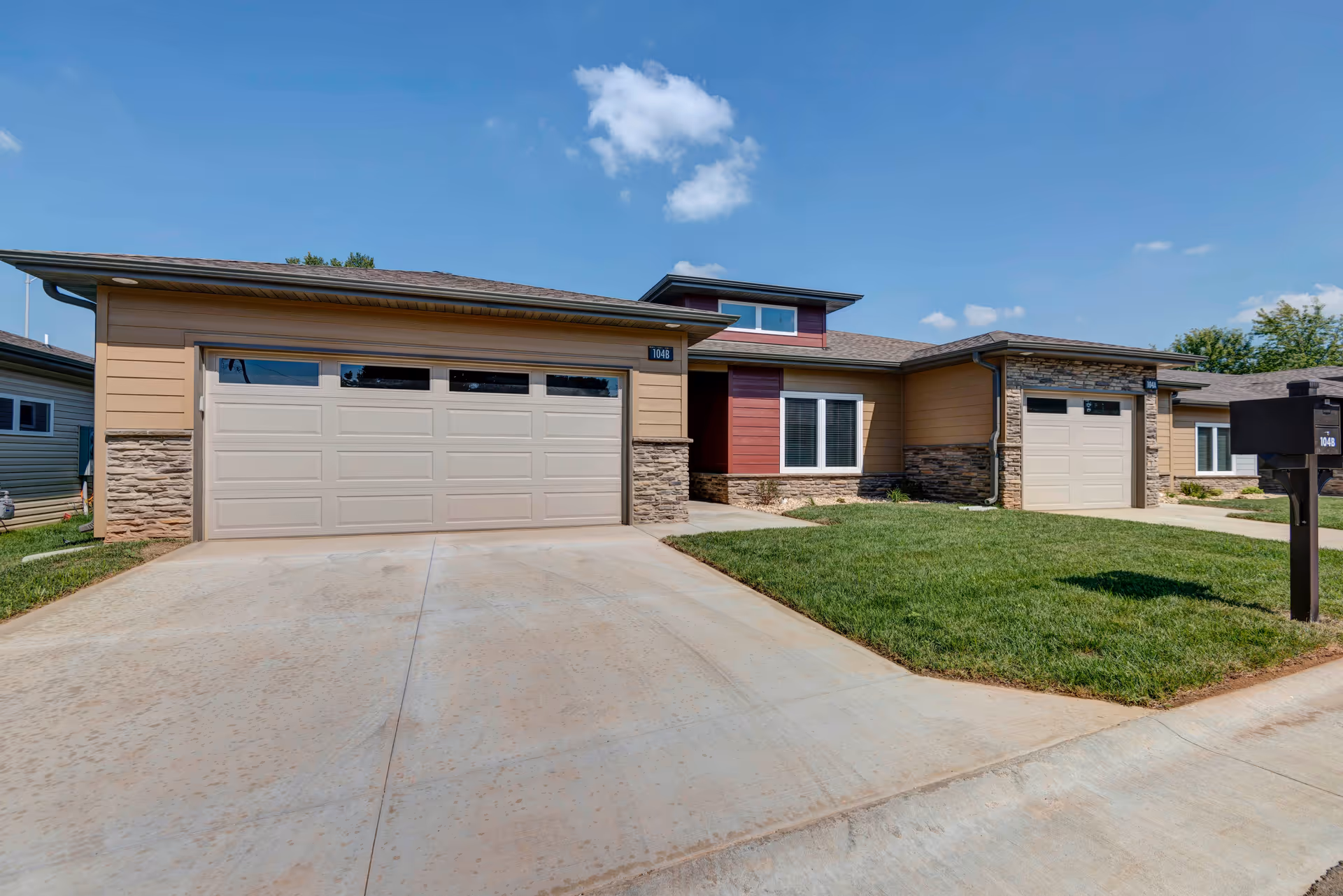 Exterior view of a single-story residential building with two beige garage doors, stone accents, and a small green lawn under a clear blue sky.
