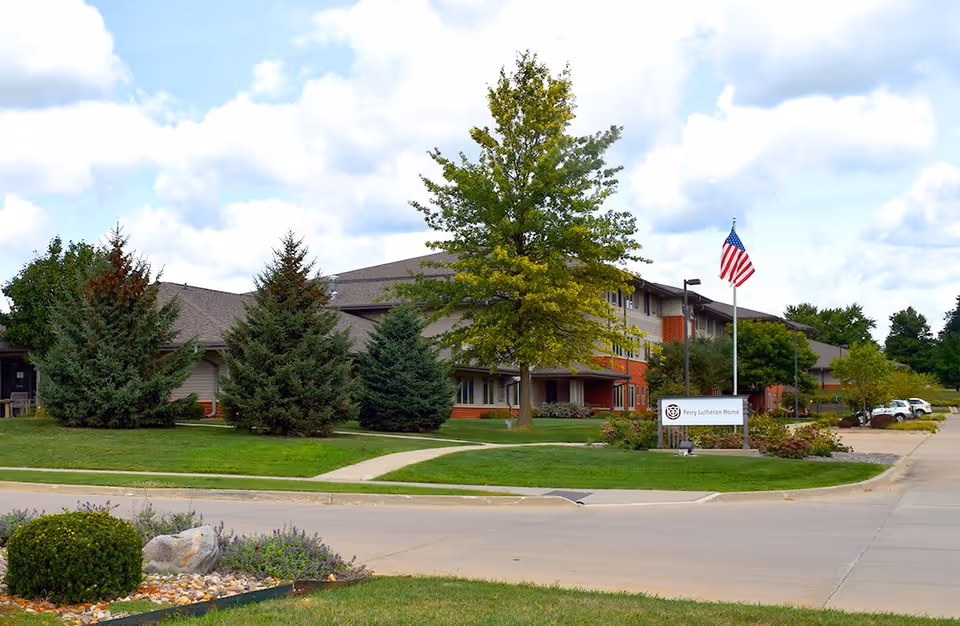 Exterior view of the Spring Valley Campus of Perry Lutheran Homes showing a multi-story building with a sloped roof, surrounded by green trees and a well-maintained lawn. An American flag is flying on a flagpole near a sign that reads 'Perry Lutheran Home'. The sky is partly cloudy.
