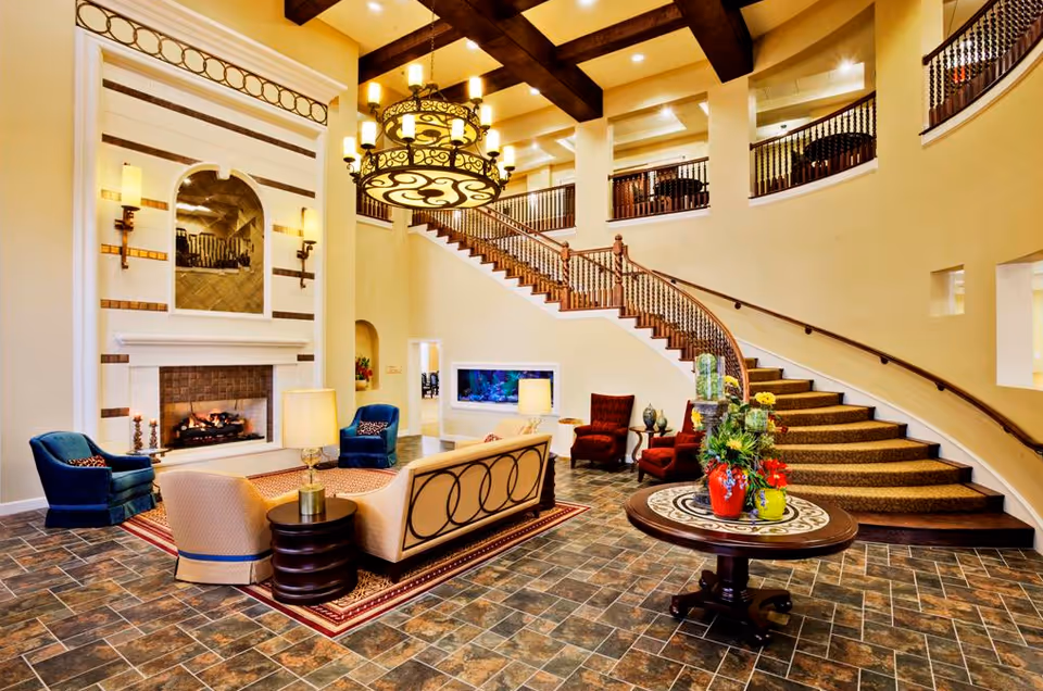 Spacious and elegant living room area with a large fireplace, comfortable seating including armchairs and a sofa, a round table with floral arrangements, and a grand curved staircase leading to an upper level with a balcony railing. The room features warm lighting from a large chandelier and wall sconces, with tiled flooring and high ceilings with exposed wooden beams.