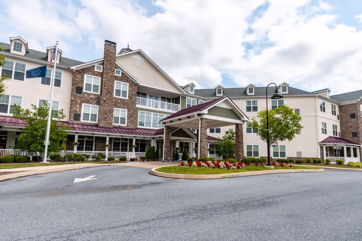 Exterior view of a multi-story senior living facility with a covered entrance, stone and beige siding, multiple windows, a flagpole with two flags, landscaped flower beds, and a curved driveway.