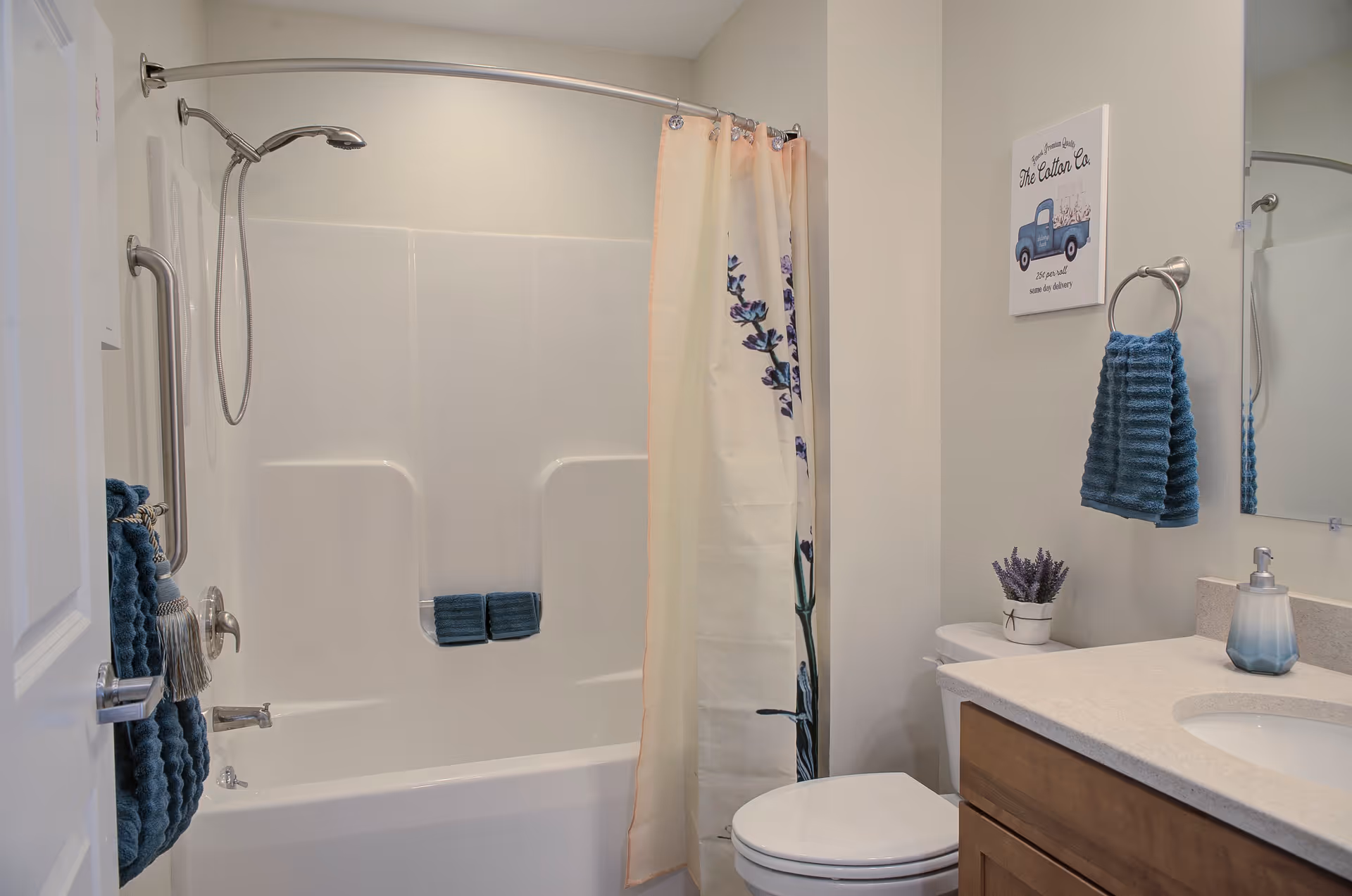 A clean bathroom featuring a white bathtub with a curved shower rod and a floral shower curtain. There are blue towels hanging on a towel rack and inside the bathtub. A toilet is next to the bathtub, and a countertop with a sink, soap dispenser, and a small potted plant is visible. A decorative wall sign and a mirror are above the sink.