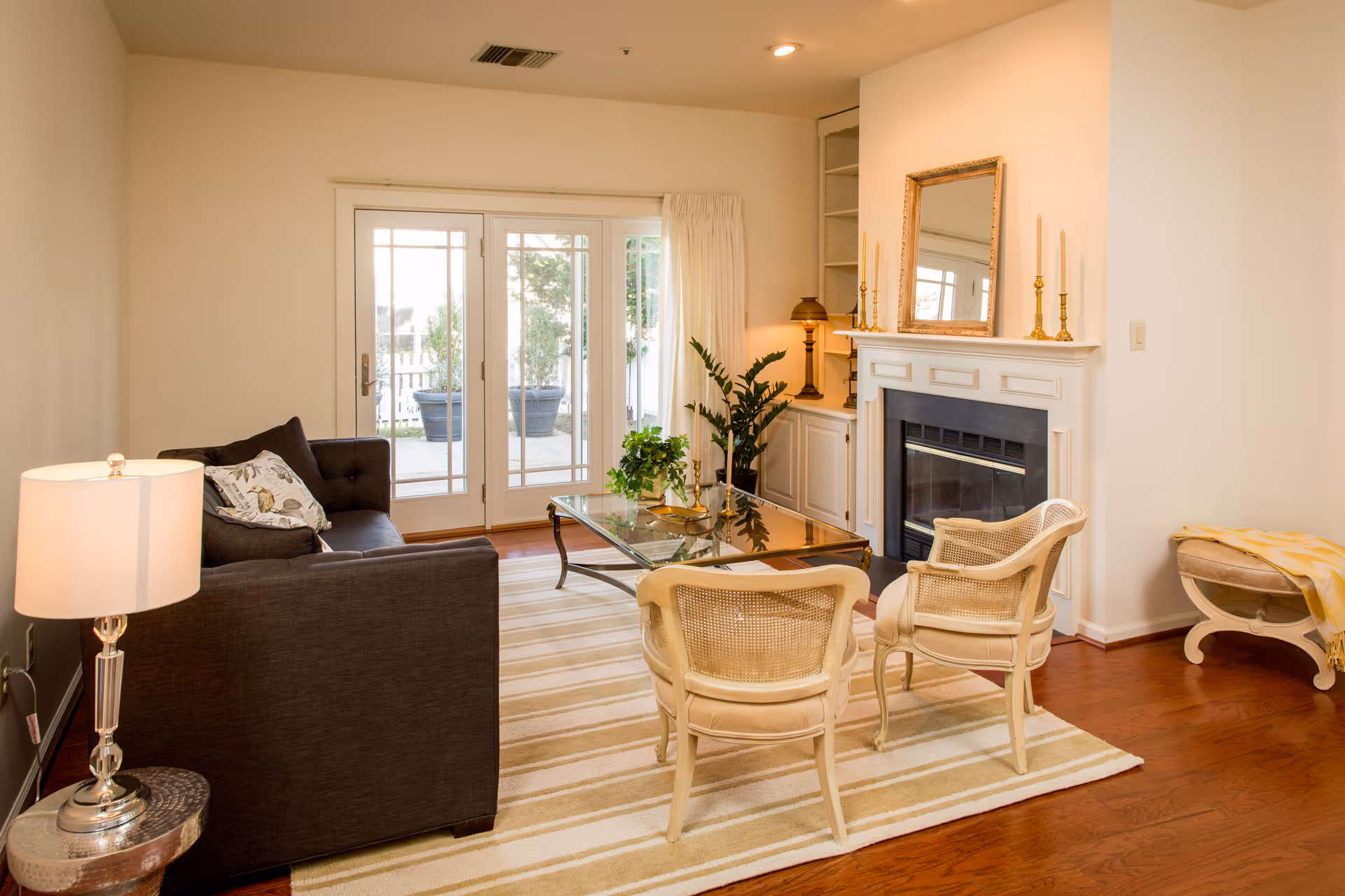 A cozy living room with a dark gray sofa, two beige cane-back chairs, a glass coffee table, and a white fireplace with a gold-framed mirror and candlesticks on the mantel. There is a lamp on a small round side table next to the sofa, a potted plant near the fireplace, and French doors leading to an outdoor area with potted plants. The room has wooden flooring and a striped area rug.