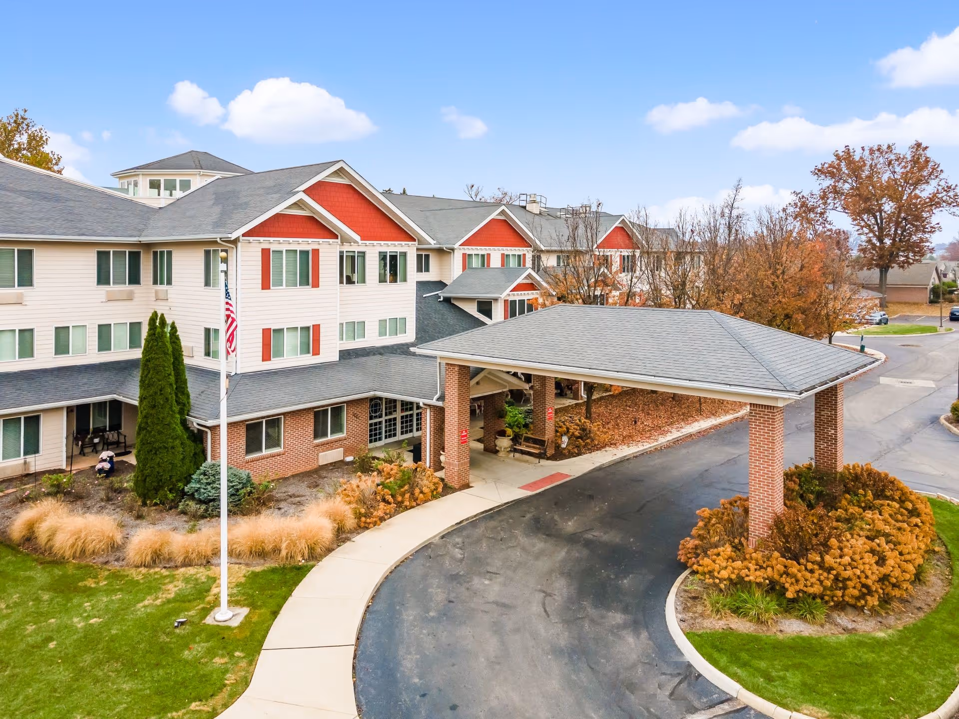 Exterior view of Rittenhouse Village Gahanna, a multi-story senior living facility with beige and red siding, a covered entrance supported by brick columns, landscaped greenery, and an American flag on a flagpole in front.
