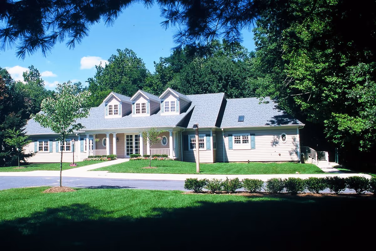 A single-story building with a gray roof and beige siding surrounded by green grass, small trees, and bushes under a clear blue sky, likely a senior living facility.