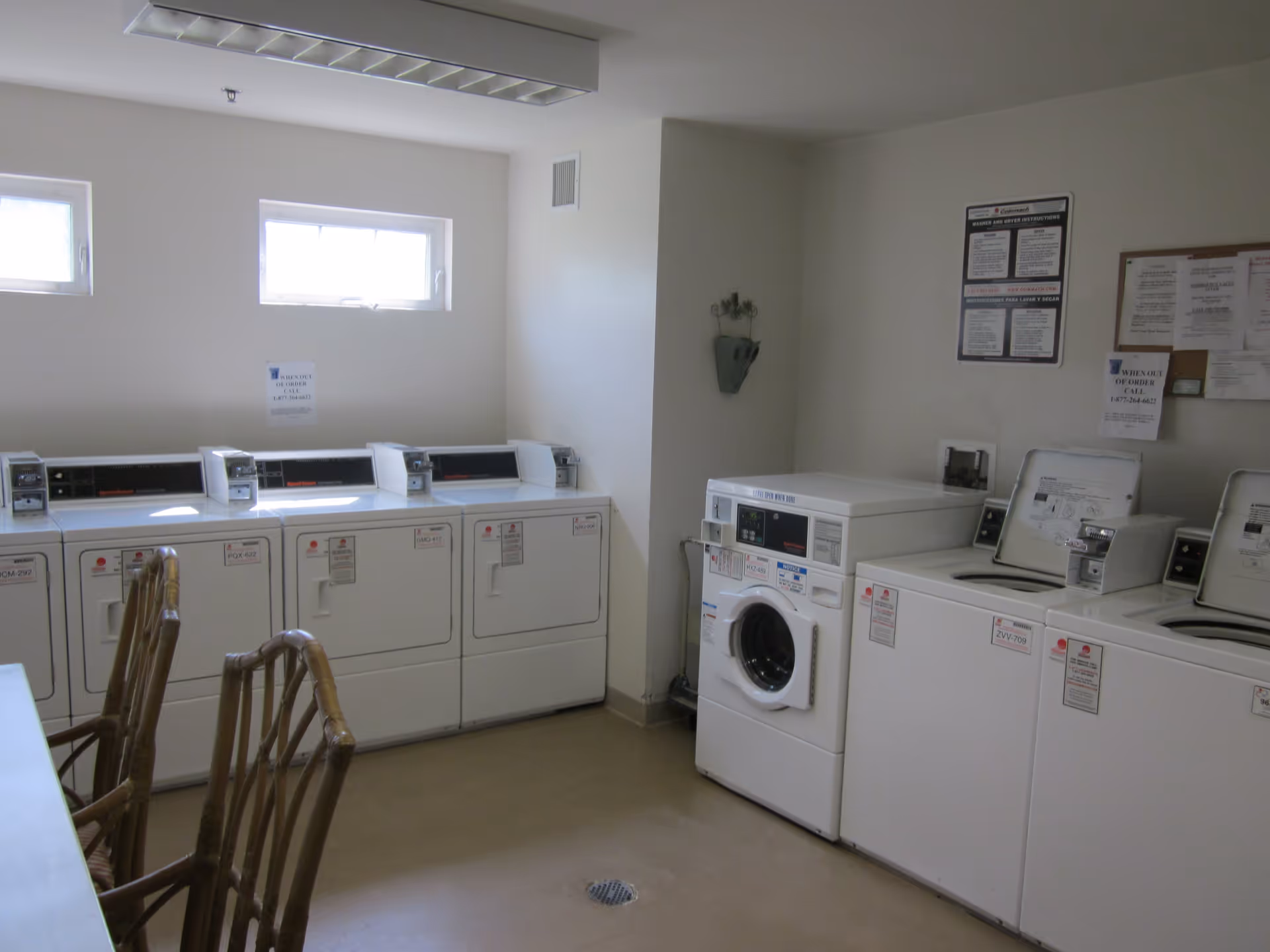 Laundry room with multiple white washing machines and dryers along the walls, a table with wooden chairs in the foreground, and windows letting in natural light.