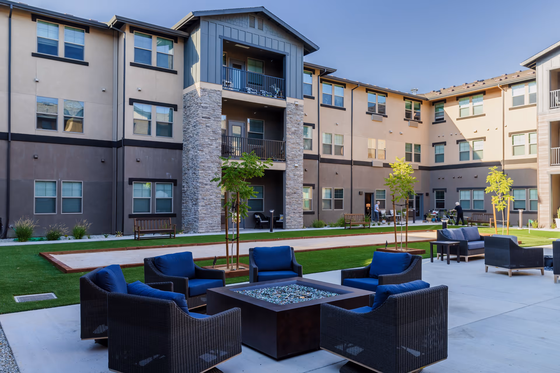 Outdoor courtyard area of a senior living facility with modern multi-story buildings surrounding a green lawn. The courtyard features several seating areas with dark wicker chairs and blue cushions arranged around a fire pit table. Small trees are planted along the lawn, and a bocce ball court is visible in the background. A few people can be seen in the distance near the building entrances.
