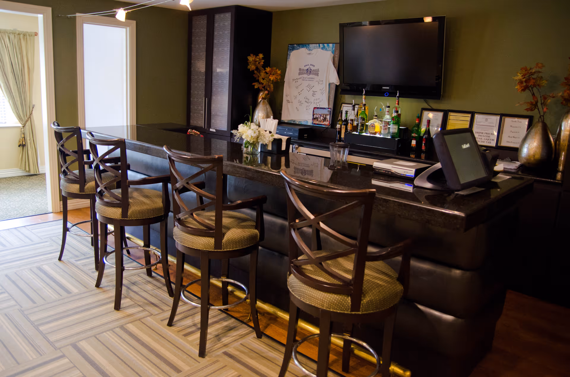 Interior view of a bar area with four wooden bar stools with cushioned seats lined up along a dark granite countertop. Behind the bar, there is a mounted flat-screen TV, various bottles of liquor, framed certificates, and decorative vases with flowers. The room has green walls and a patterned carpet floor, with an adjacent room visible through an open doorway.