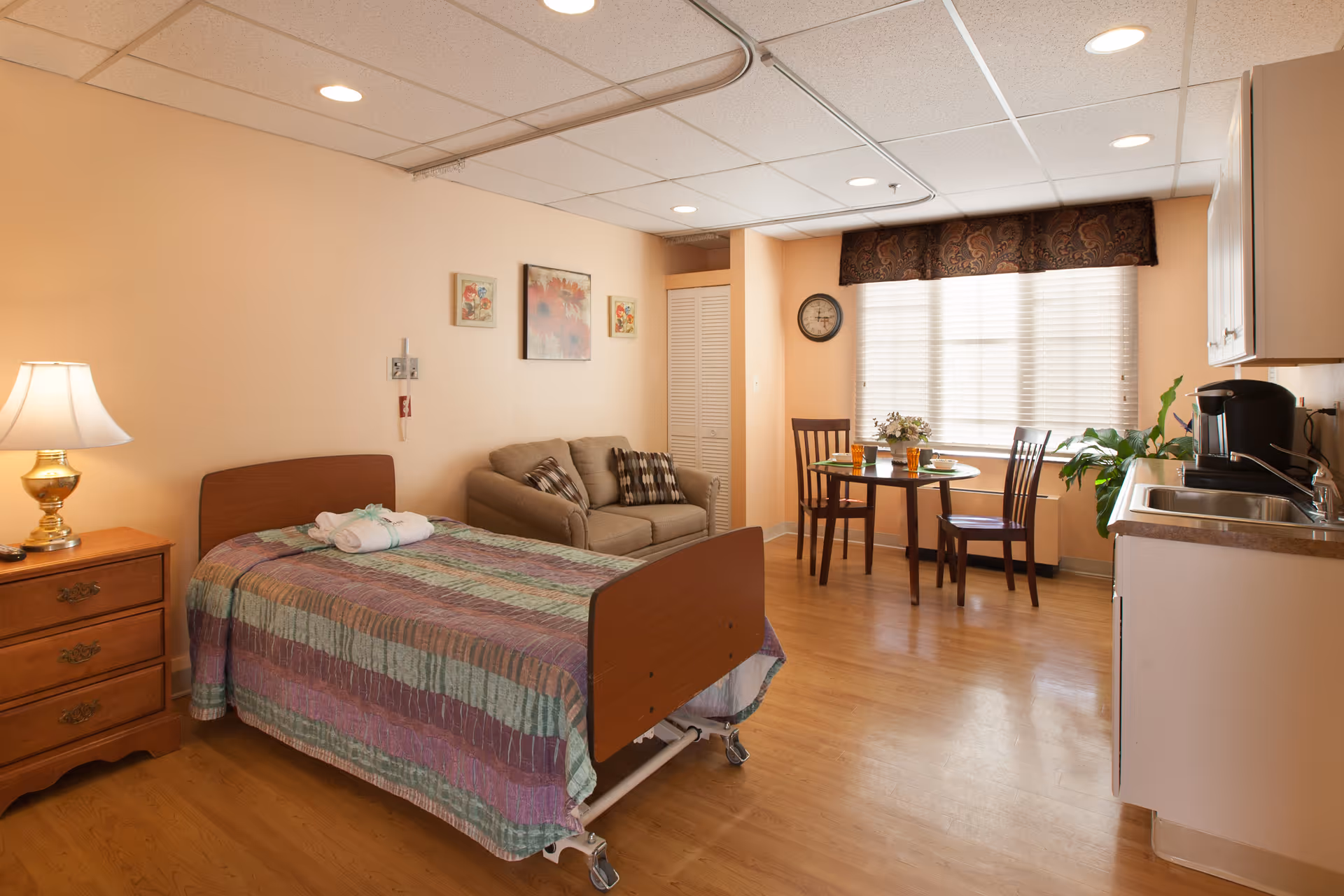 A cozy nursing home room with a single bed covered in a striped quilt, a wooden nightstand with a lamp, a small sofa with patterned pillows, a round dining table with two chairs near a window with blinds and a valance, a wall clock, and a kitchenette area with a sink and coffee maker. The room has light peach walls and wood flooring.