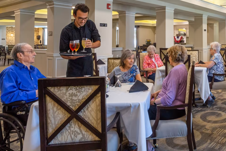 A group of elderly people sitting at tables in a dining room of a senior living facility. A waiter is serving drinks to one of the tables. The room has white tablecloths, cushioned chairs, and decorative columns.