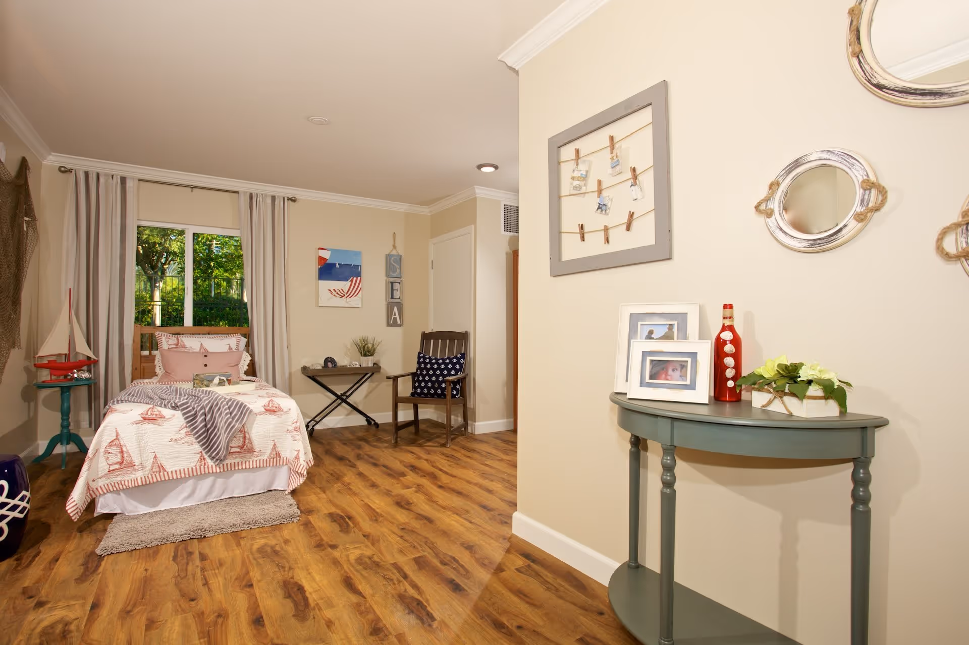 A cozy bedroom with a single bed covered in a white and red sailboat-patterned quilt, a small green side table with a decorative sailboat, a wooden chair with a navy blue pillow, and a small table with a plant and clock. The room has wooden flooring, beige walls, a window with white curtains, and nautical-themed wall decorations including a 'SEA' sign and framed photos on a green console table.