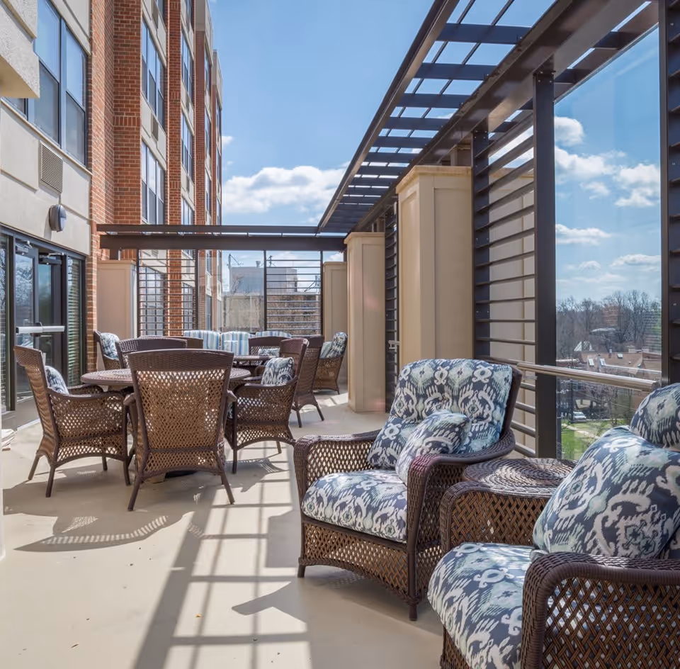 Outdoor patio area at Sunrise of Chevy Chase with wicker chairs and tables featuring patterned cushions, surrounded by glass and metal railings, adjacent to a brick building under a clear blue sky.