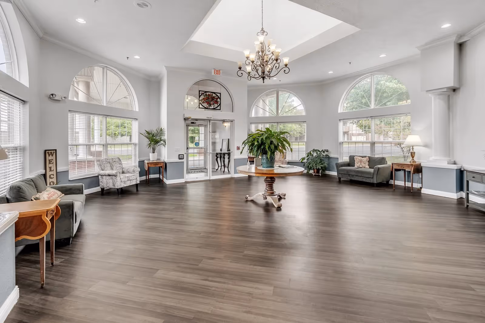 Spacious, well-lit lobby/common area with seating, large arched windows, a central round table with plants, and a chandelier.