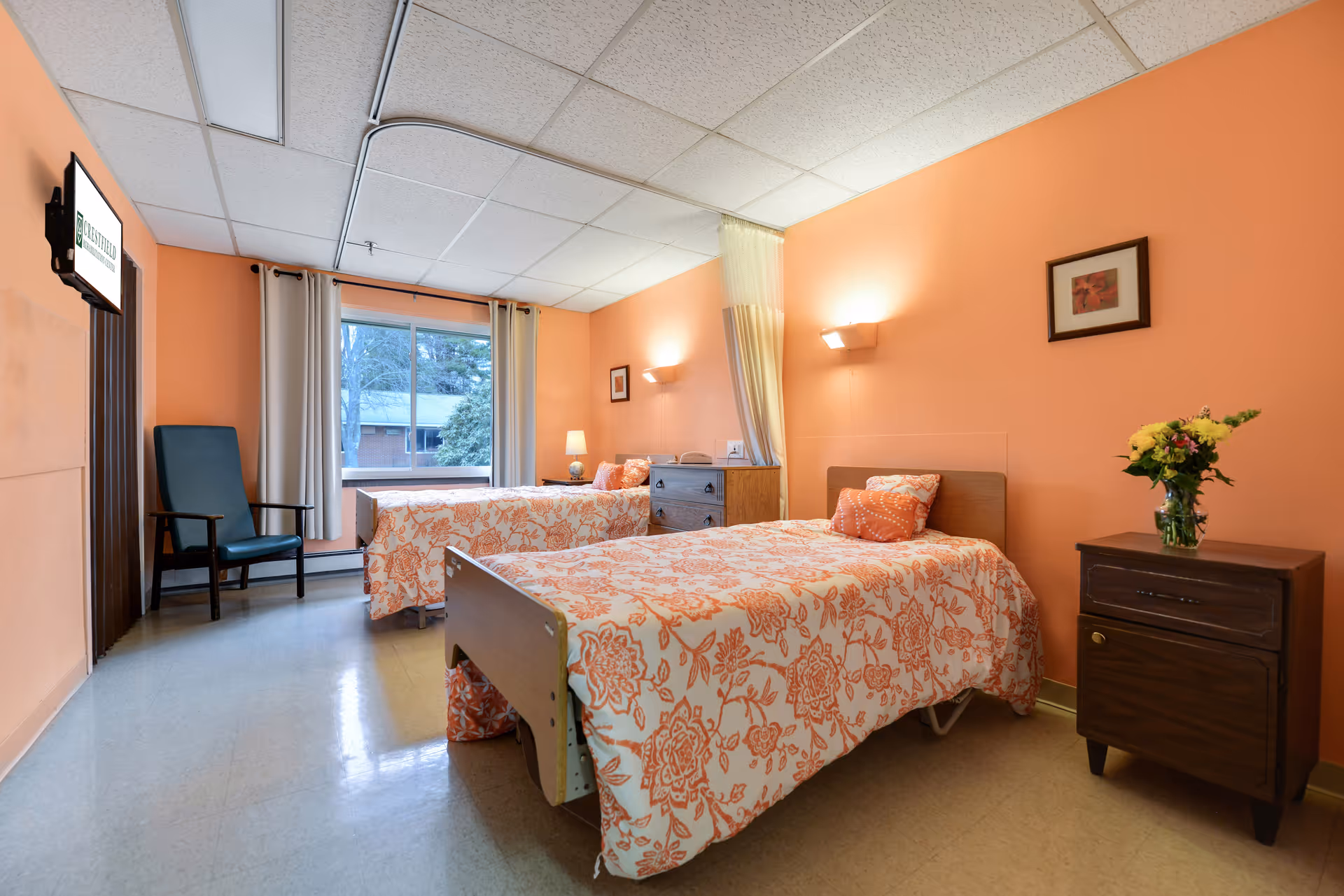 A bright and clean bedroom in a senior living facility with two single beds covered in matching orange and white floral bedspreads. The walls are painted peach, and there is a window with white curtains letting in natural light. A wooden nightstand with a vase of flowers and a framed picture on the wall add a homey touch. A green armchair is placed near the window, and a TV is mounted on the wall opposite the beds.