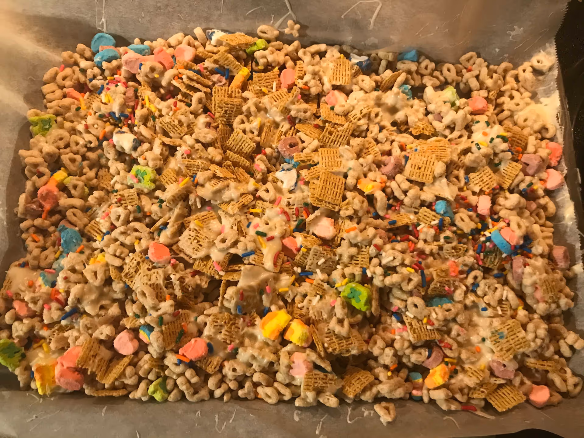Pan of cereal snack mix with marshmallows, Chex pieces, Cheerios and sprinkles on parchment paper.