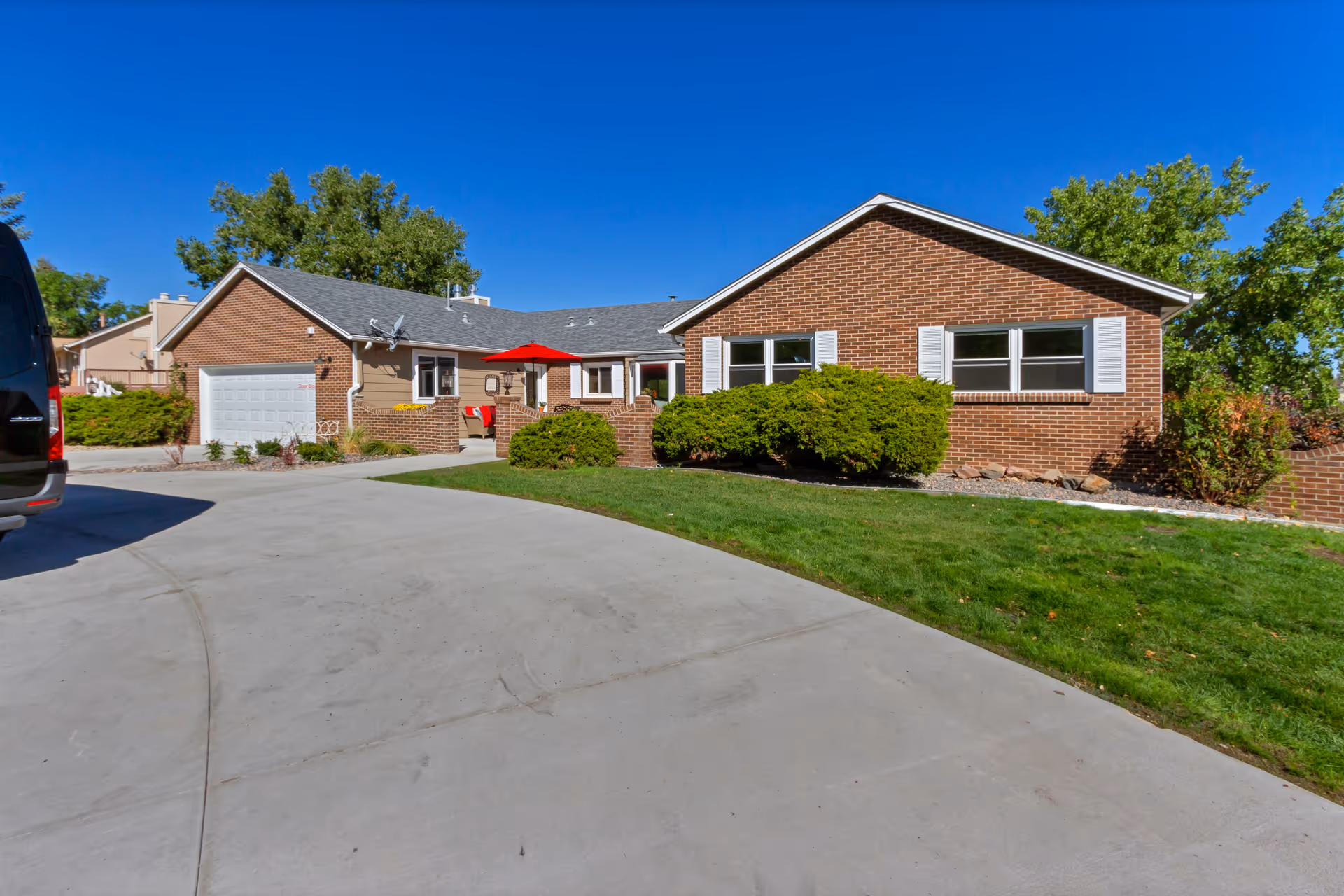 Exterior view of a single-story brick building with white window shutters, a garage, a concrete driveway, green lawn, bushes, and a red patio umbrella under a clear blue sky.