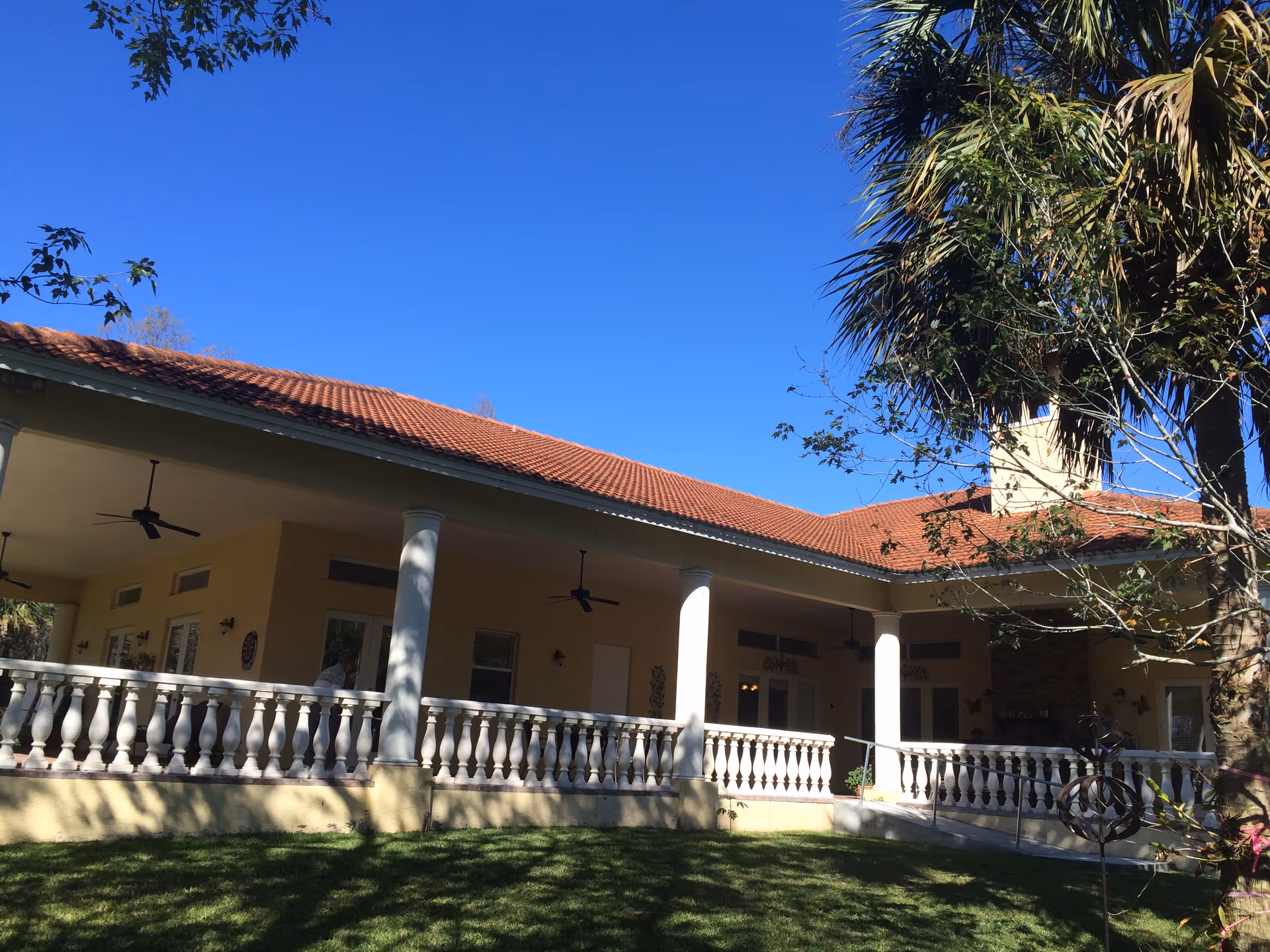 Exterior view of a single-story building with a red tiled roof and a covered porch supported by white columns. The porch has ceiling fans and a white balustrade. There is a green lawn in the foreground and trees on the right side under a clear blue sky.