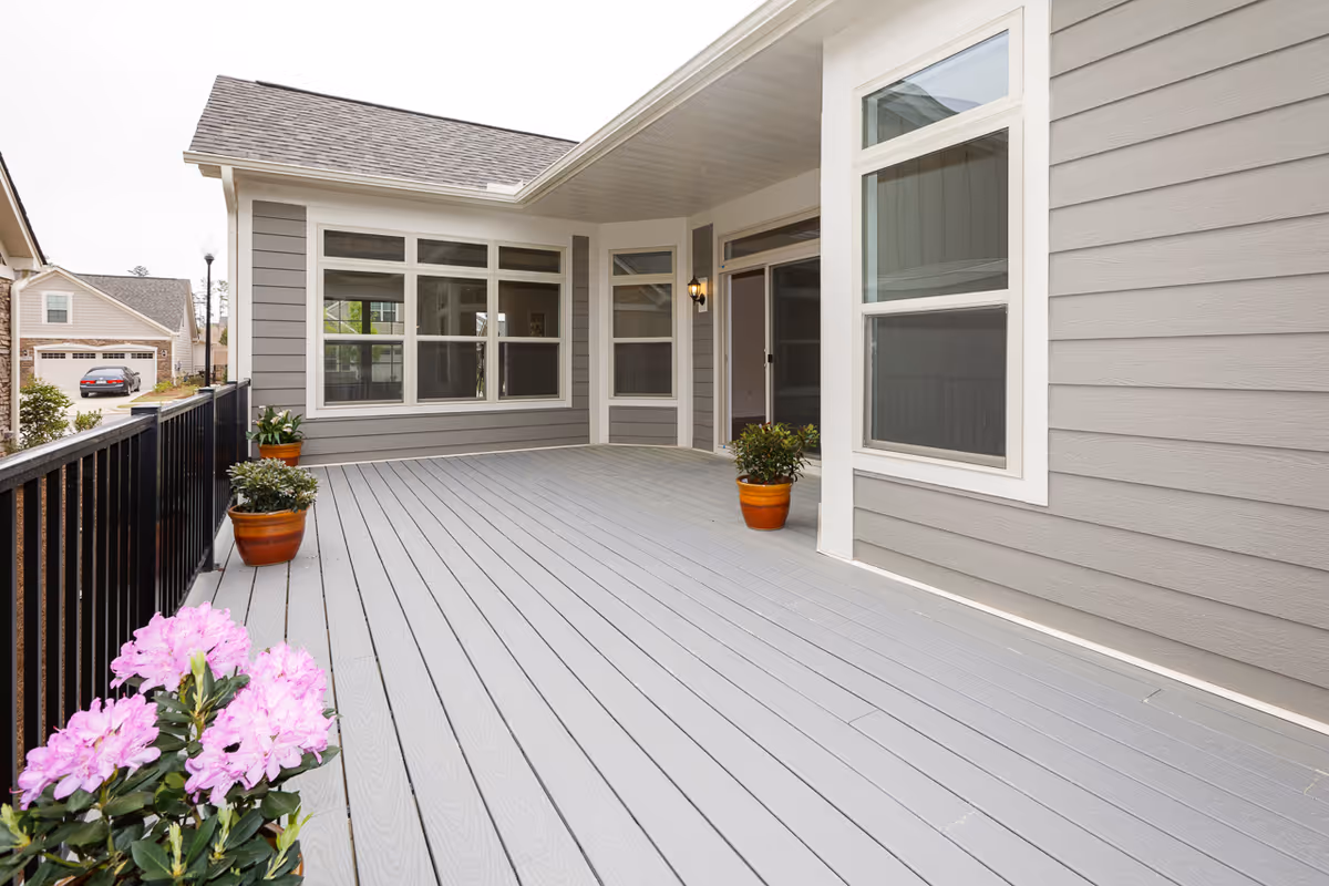 A spacious outdoor patio area with gray wooden flooring and light gray siding walls. The patio is partially covered with a white ceiling and features several potted plants, including pink flowers in the foreground and green plants in terracotta pots along the edges. There is a black metal railing on one side and multiple windows and a sliding glass door leading into the building.