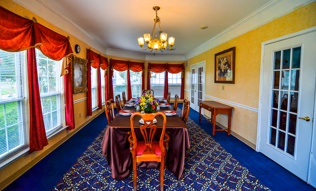 A formal dining room with a long table covered with a dark tablecloth, set with plates and napkins. The room has large windows with red curtains and valances, a blue patterned carpet, yellow walls, a chandelier hanging from the ceiling, and a framed painting on the wall. There are wooden chairs around the table and a small wooden side table against the wall.