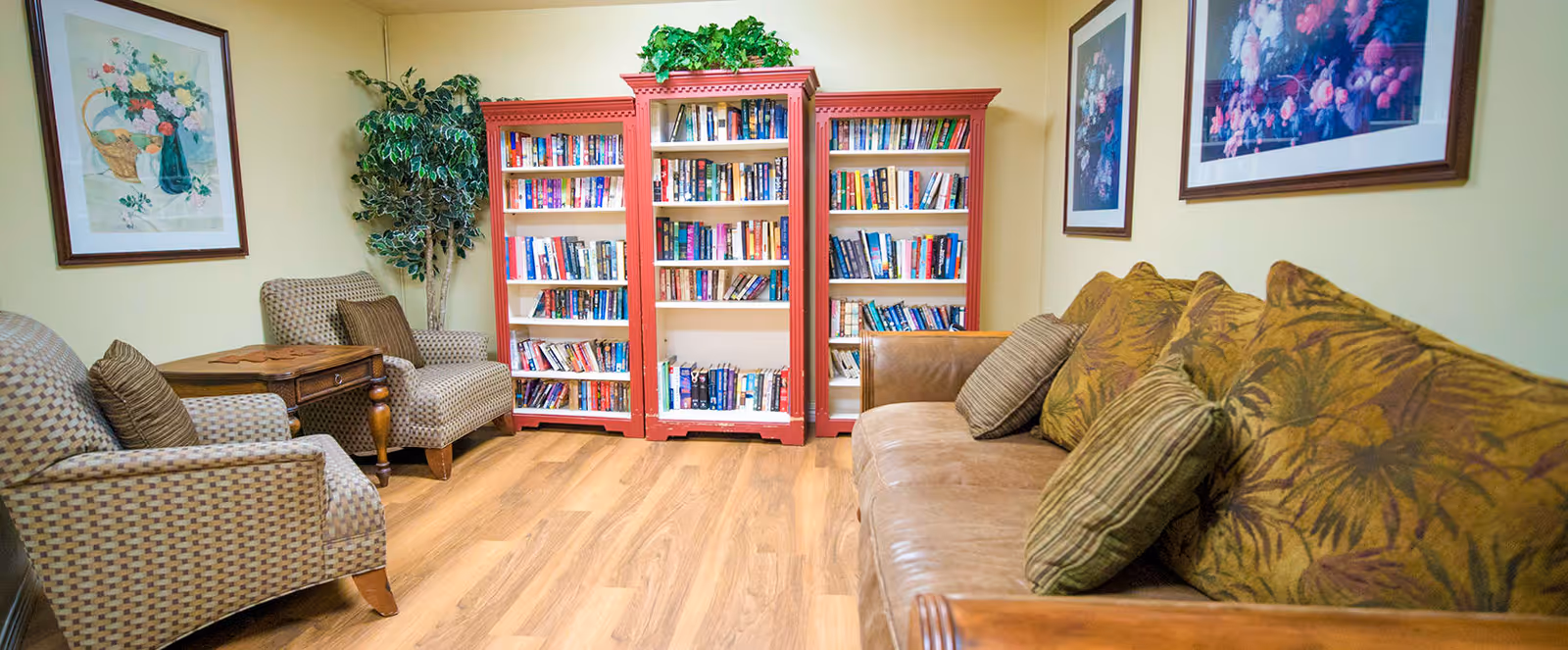 A cozy living room area with a brown leather sofa adorned with patterned cushions on the right, two upholstered armchairs with a small wooden side table between them on the left, a red bookshelf filled with books against the back wall, a potted plant next to the bookshelf, and framed floral artwork hanging on the walls.