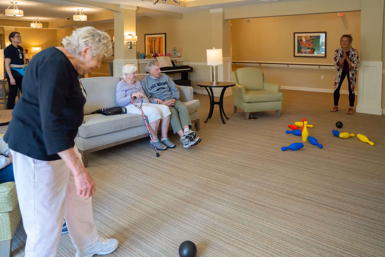 A group of elderly people and a staff member are in a spacious, well-lit common area with beige walls and carpeted floor. Two elderly individuals are seated on a beige couch, one holding a cane, while another elderly woman is standing and rolling a black ball on the floor towards a set of colorful plastic bowling pins. Another woman stands near the pins, watching the game. The room is furnished with a green armchair, a round side table with a lamp, a piano, and wall art.