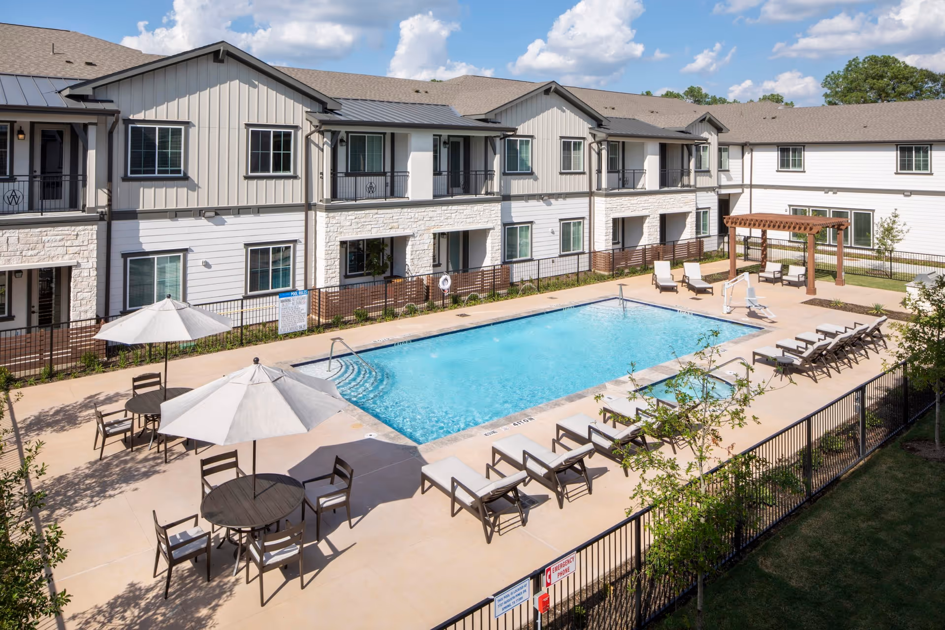 Outdoor swimming pool area at The Landing at Augusta Woods Senior Living with lounge chairs, tables with umbrellas, and a pergola. The pool is surrounded by a fence and the senior living building is visible in the background under a partly cloudy sky.