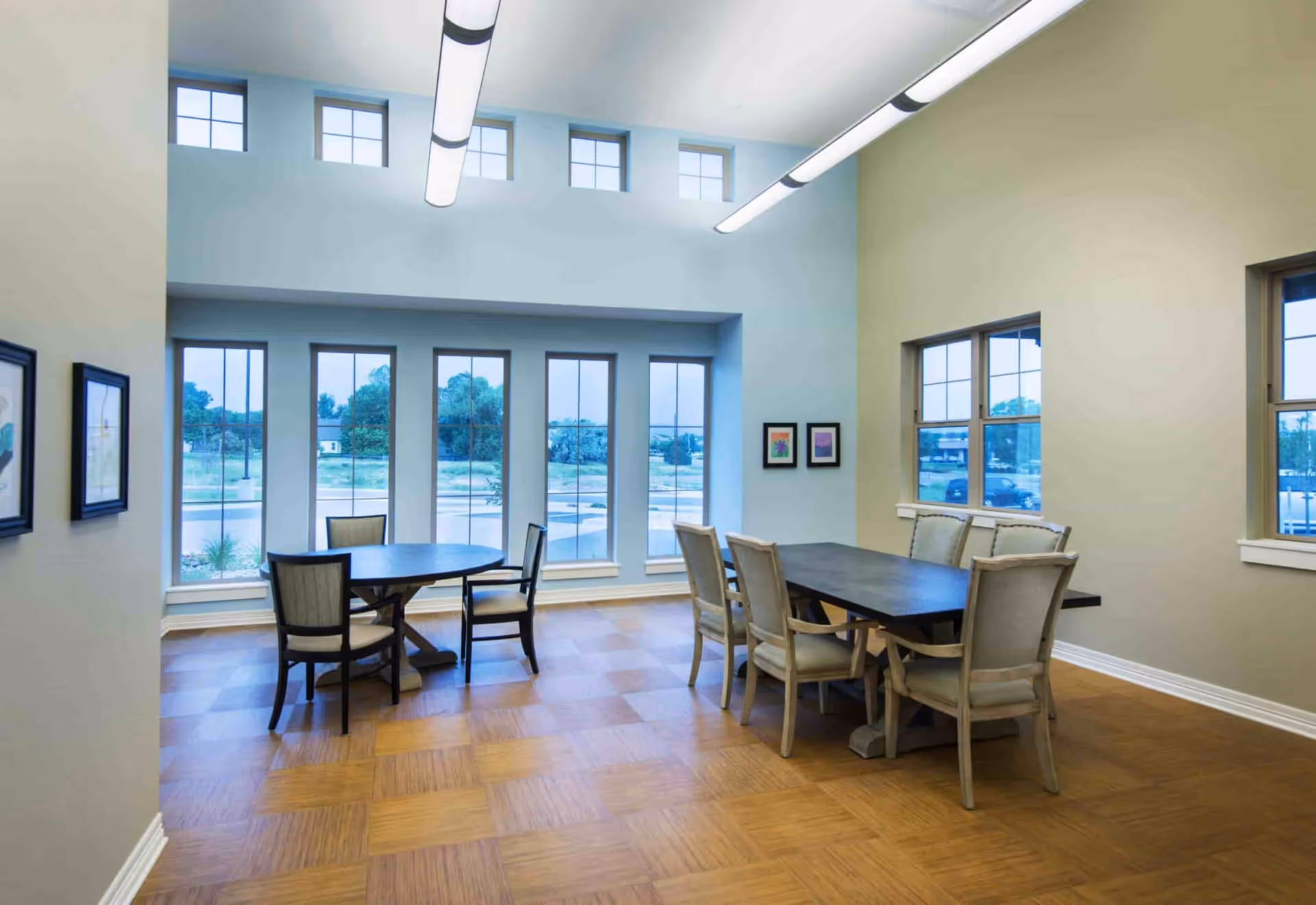 A bright dining room with large windows letting in natural light. The room features two tables, one round with four chairs and one rectangular with six chairs. The walls are painted in soft pastel colors, and there are framed pictures hanging on the walls. The floor has a wood pattern, and modern ceiling lights illuminate the space.