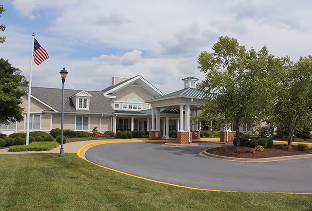 Front entrance of a single-story assisted living facility with a covered porte-cochère, circular driveway, flagpole, and landscaped grounds.