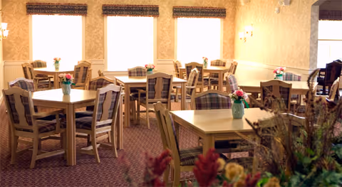 A bright dining room with multiple wooden tables and chairs featuring plaid cushions. Each table has a small vase with pink flowers. The room has large windows with woven blinds, beige patterned wallpaper, and wall sconces providing soft lighting.