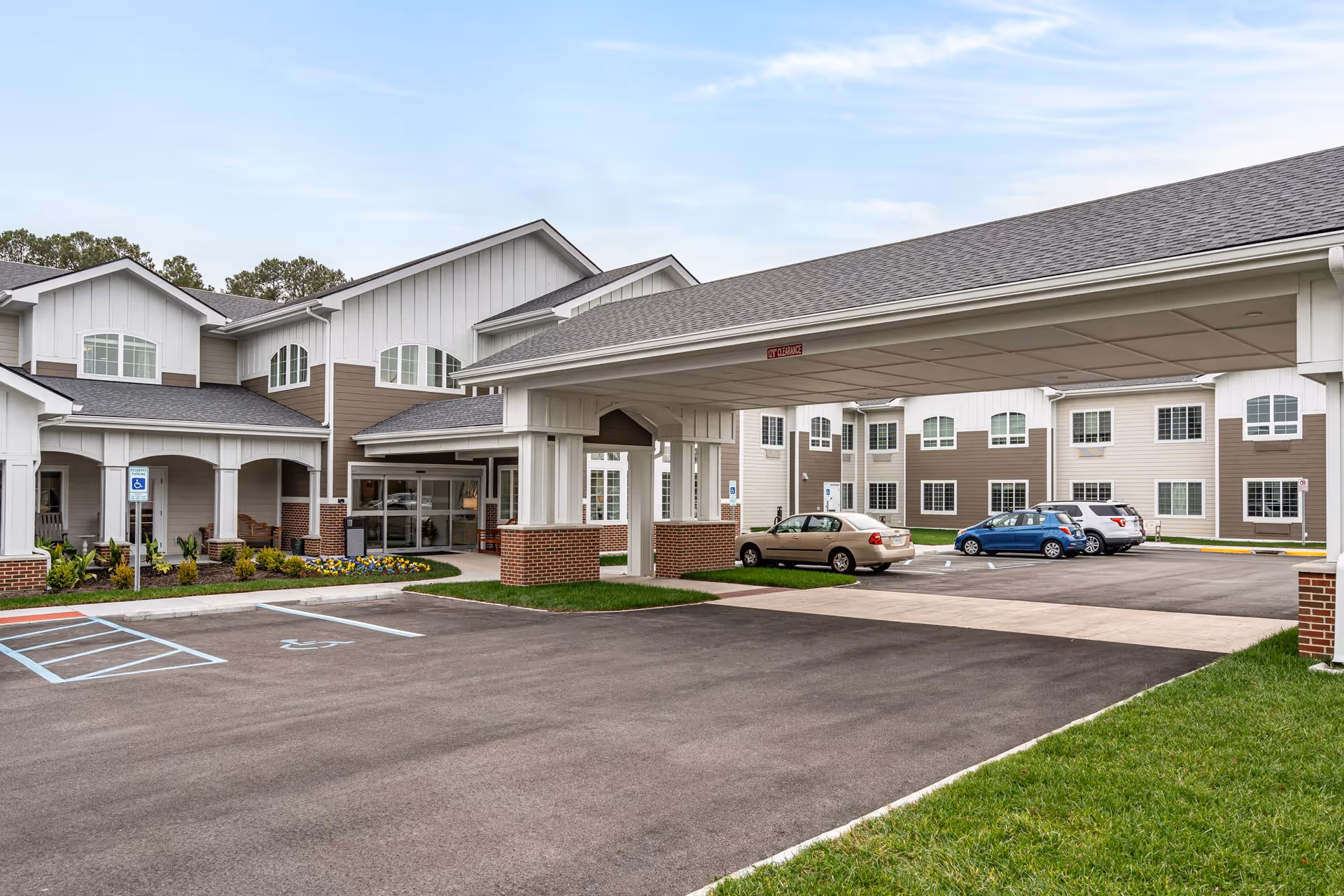 Front entrance of a senior living facility with a covered drop-off canopy, parking spaces and a multi-story building in the background.