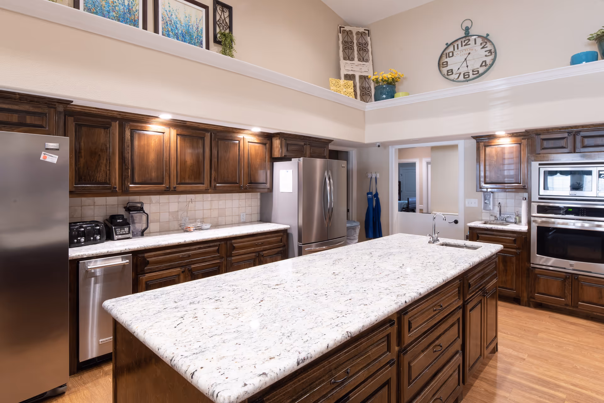 A spacious kitchen with dark wooden cabinets, a large white granite island countertop with a built-in sink, stainless steel refrigerator, dishwasher, microwave, and oven. The kitchen has a high ceiling with decorative items and a large wall clock above the cabinets.