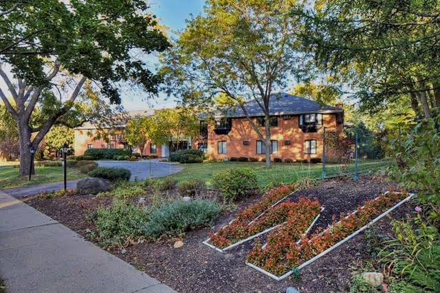 A landscaped outdoor area at Whitcomb Square Apartments featuring a garden bed shaped like the letter 'W' filled with red flowers, surrounded by trees and shrubs, with a brick apartment building in the background under a clear blue sky.